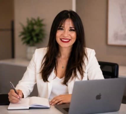 Smiling woman in white blazer sitting at a desk with a laptop and open notebook.